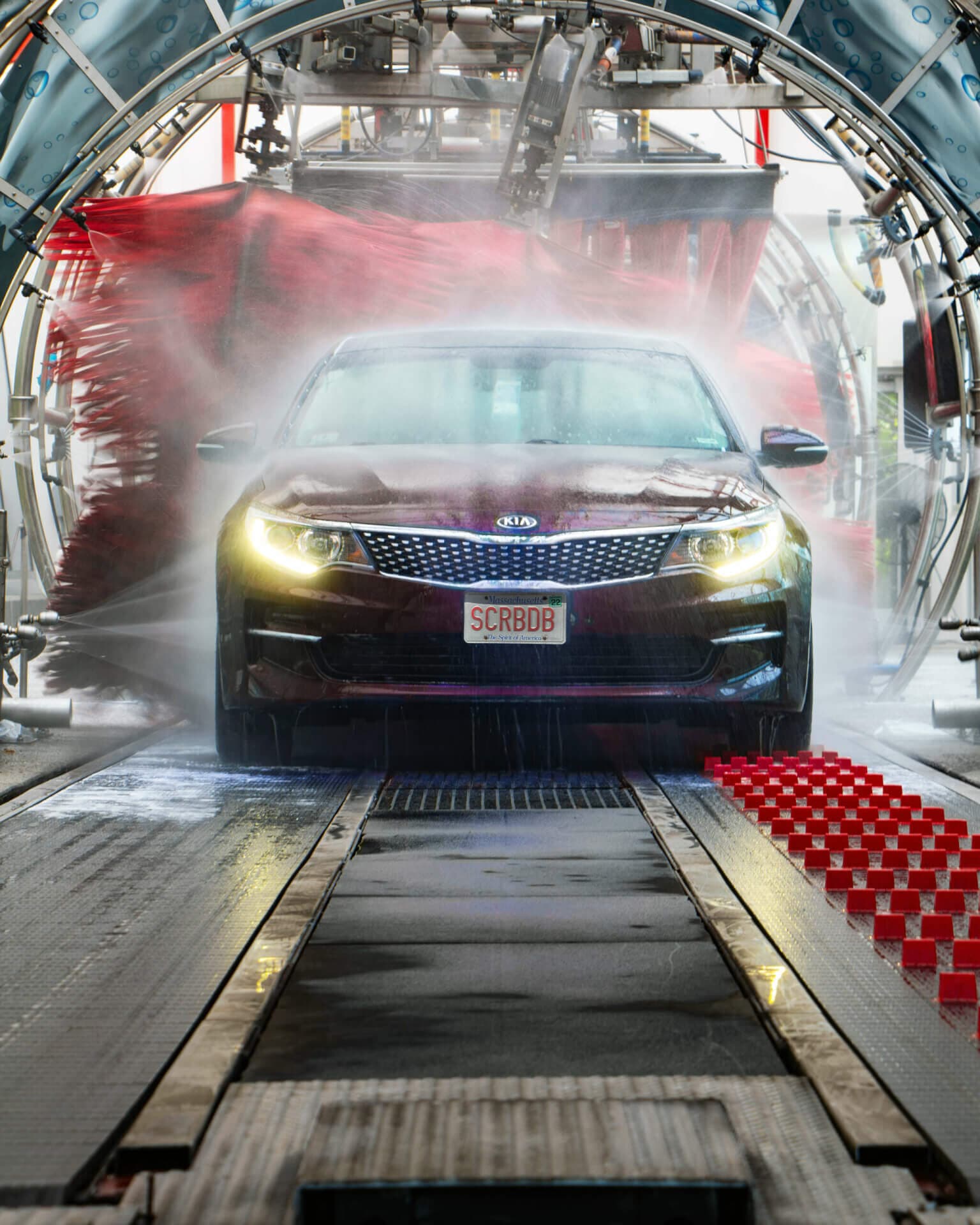Sedan with headlights on rolling head-on through a mist of rinse water between red brushes