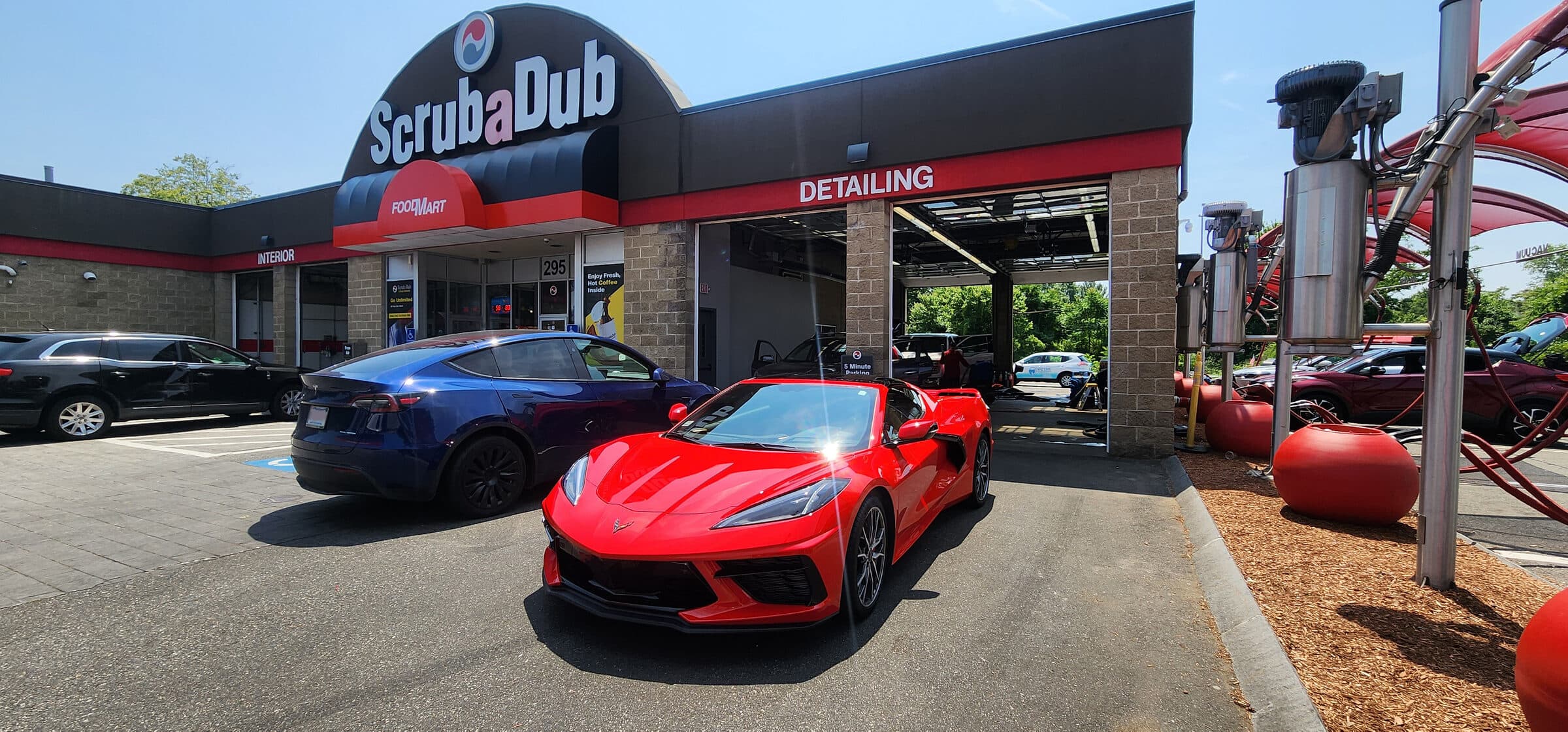 Red Corvette and blue Tesla parked in front of the ScrubaDub Woburn building with its FoodMart and Detailing bays under a clear sky
