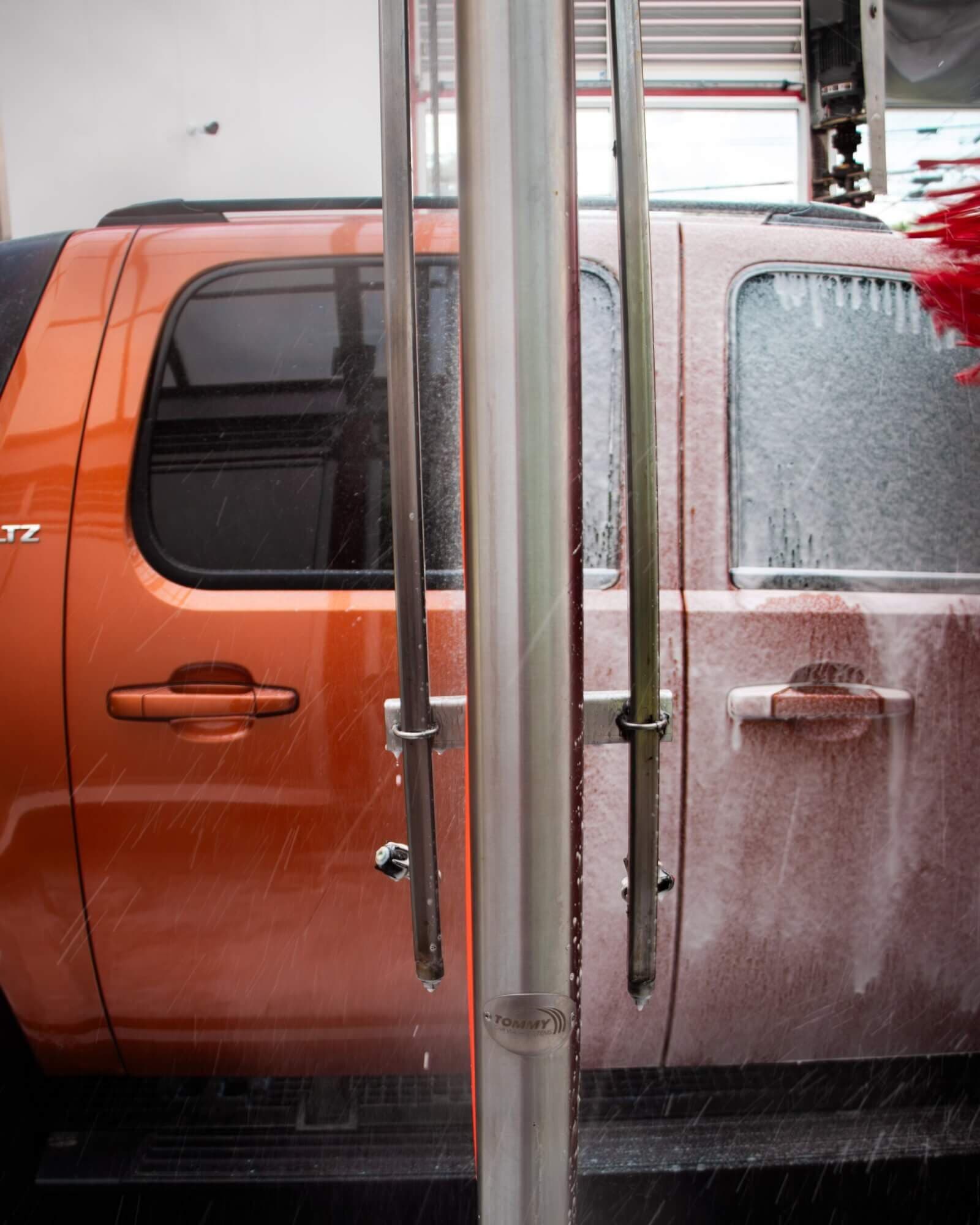 Orange pickup truck half covered in foam passing stainless steel spray arches in the wash tunnel