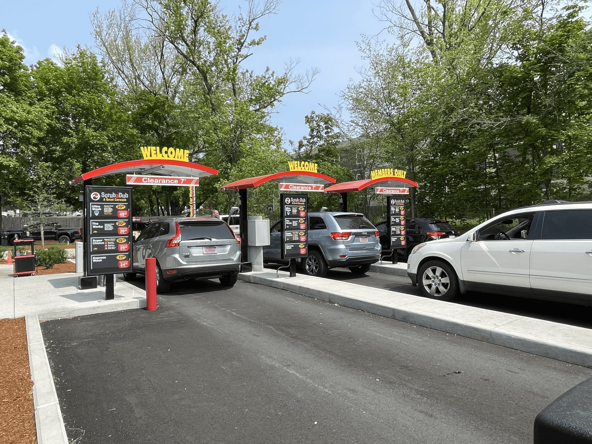 Cars lined up under three red Welcome and Members Only canopies with wash menu boards at ScrubaDub Roslindale