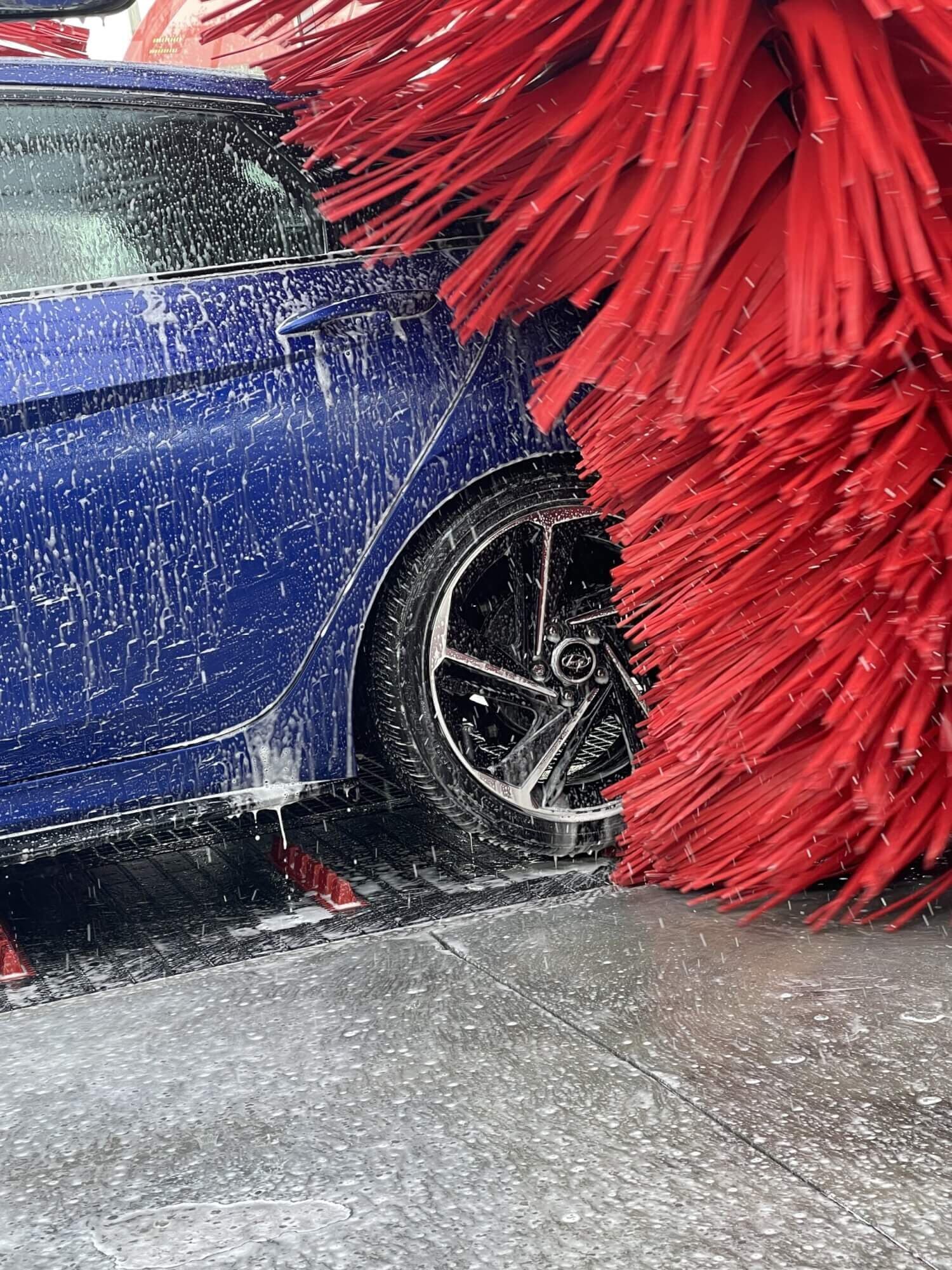Red wash brushes scrubbing a soapy blue sedan near its rear wheel