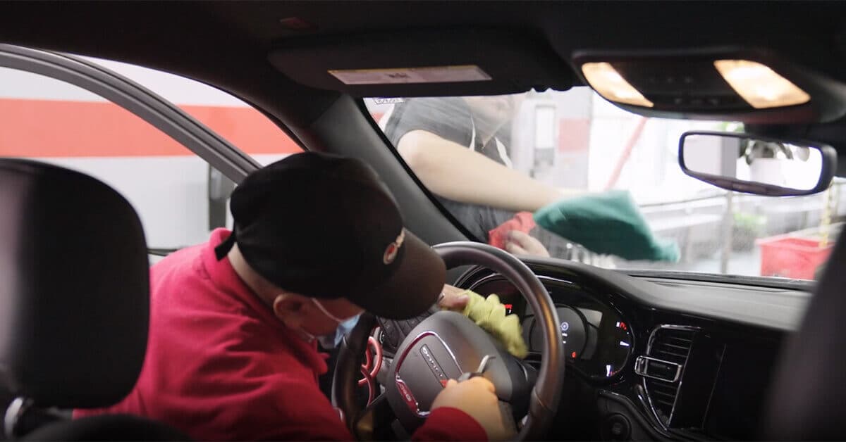 ScrubaDub attendant in a red hoodie and cap wiping a steering wheel while a coworker cleans the windshield from outside