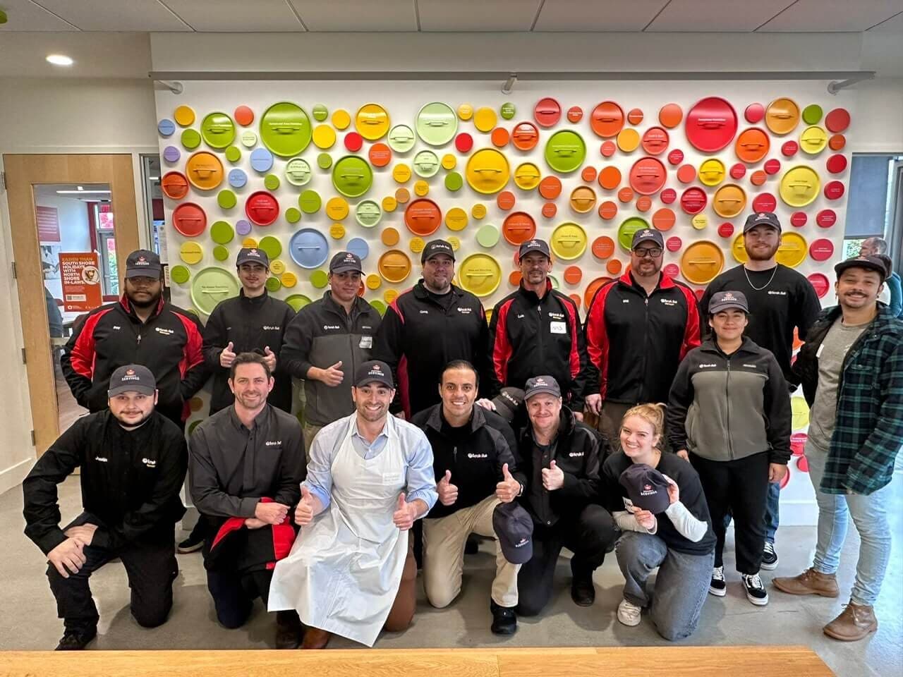 Large group of ScrubaDub employees giving thumbs up in front of a colorful wall of pot lids at Community Servings