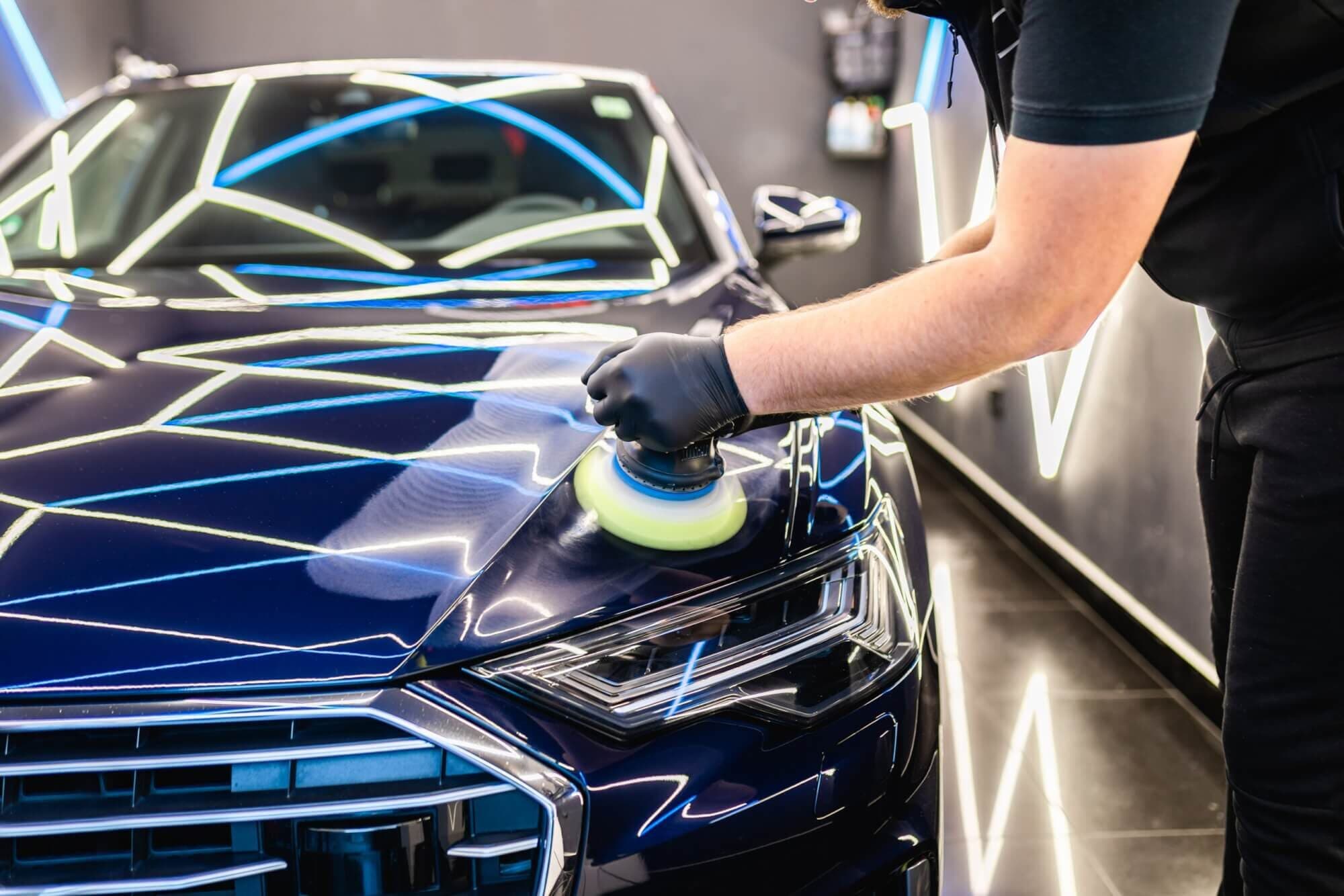 Detailer machine-polishing the hood of a dark blue sedan under geometric LED lights