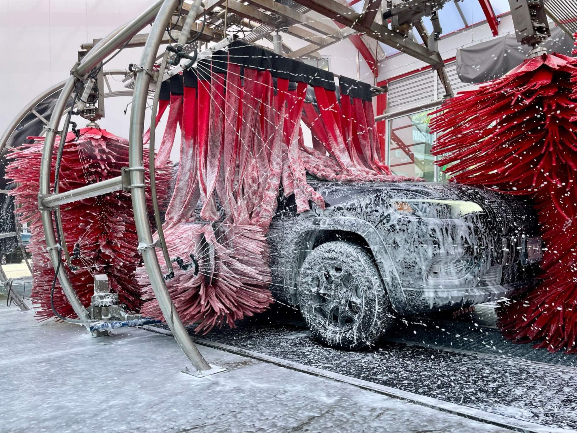 Foam-covered black SUV surrounded by red cloth curtain strips and spinning red brushes in a bright tunnel