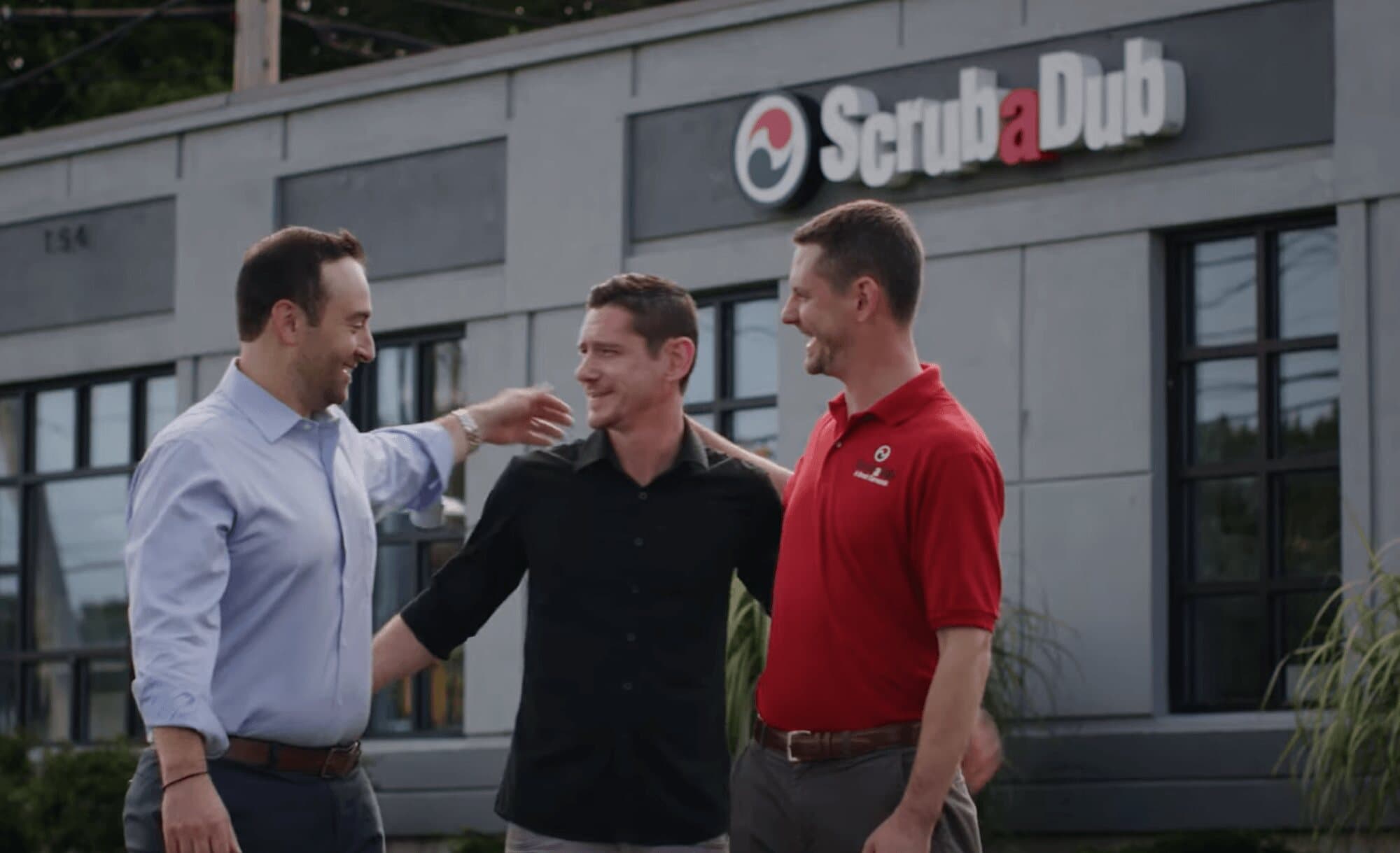 Three men laughing and embracing in front of a ScrubaDub building sign
