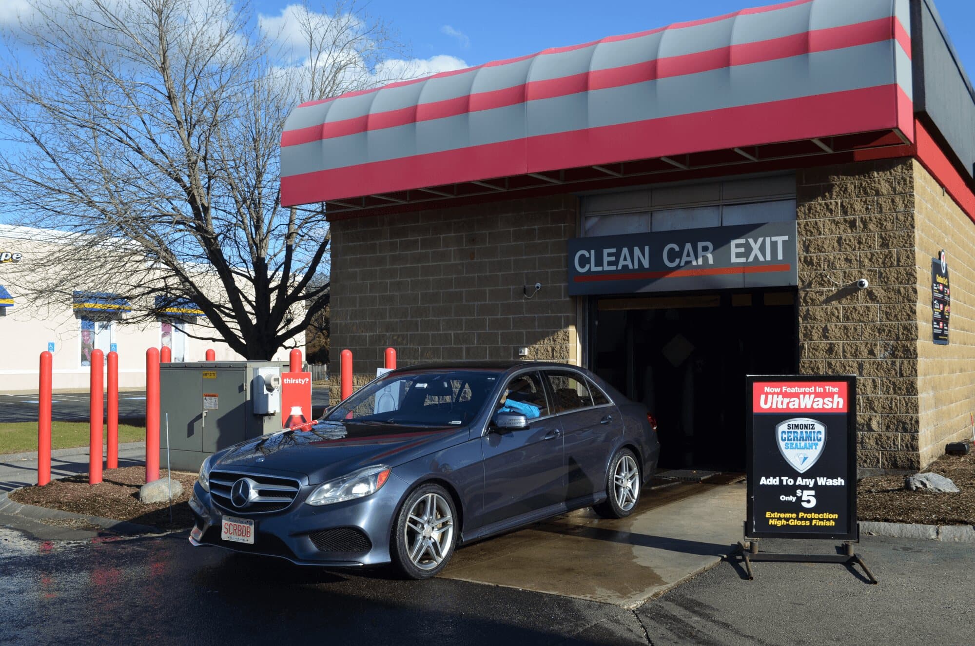 Gray sedan with a SCRBDB plate driving out of a Clean Car Exit beside an UltraWash ceramic sealant sign