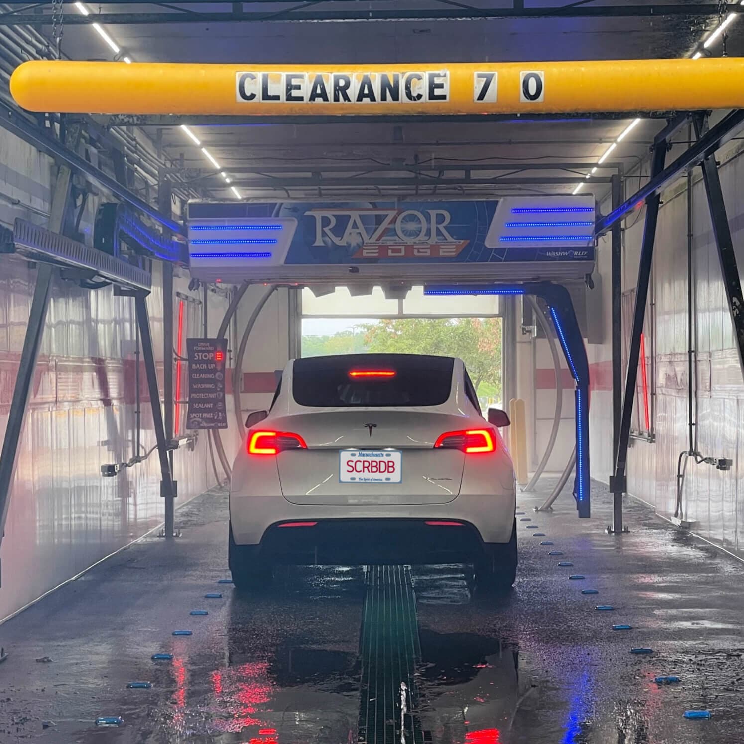 White Tesla with a SCRBDB Massachusetts plate inside a touchless in-bay car wash under a yellow 7-foot clearance bar