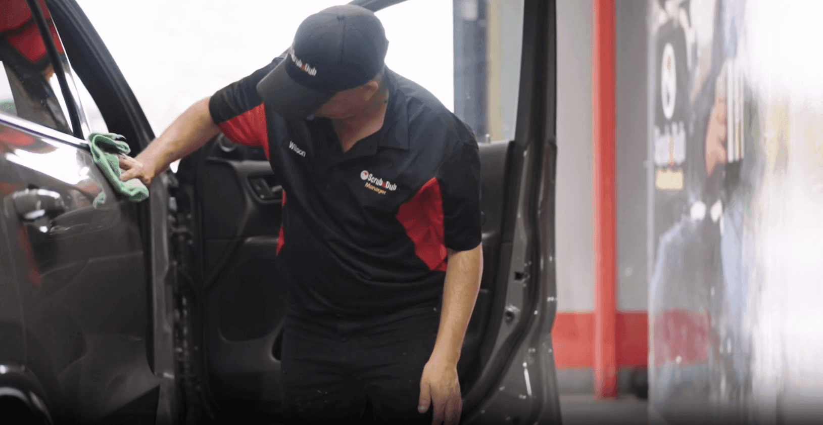 ScrubaDub manager in a black-and-red uniform and cap wiping a car door with a green microfiber towel