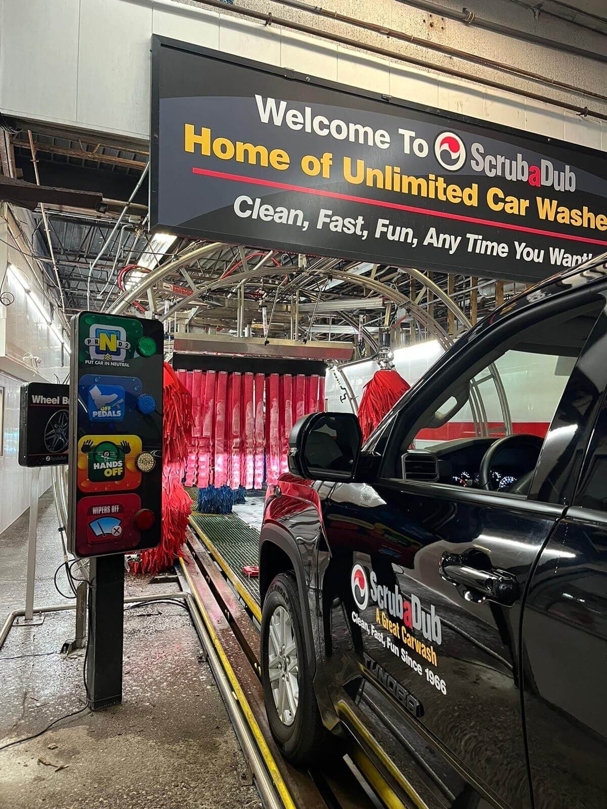 ScrubaDub-branded black pickup at the tunnel entrance under a Welcome to ScrubaDub, Home of Unlimited Car Washes sign