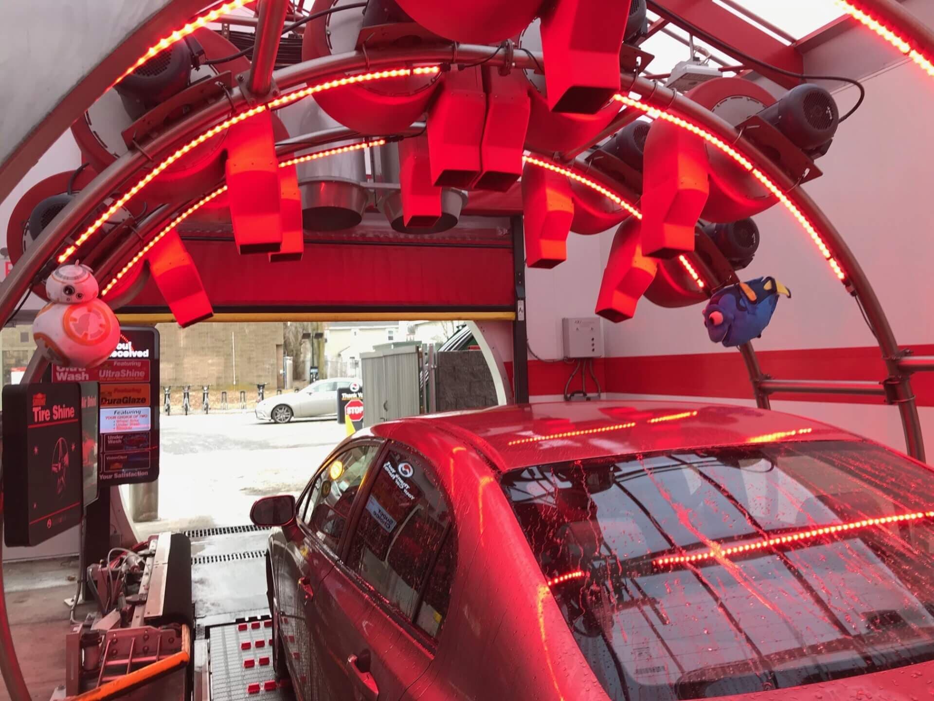 Wet red car exiting a car wash tunnel under red-lit dryers, with toy BB-8 and Dory figures hanging from the arch