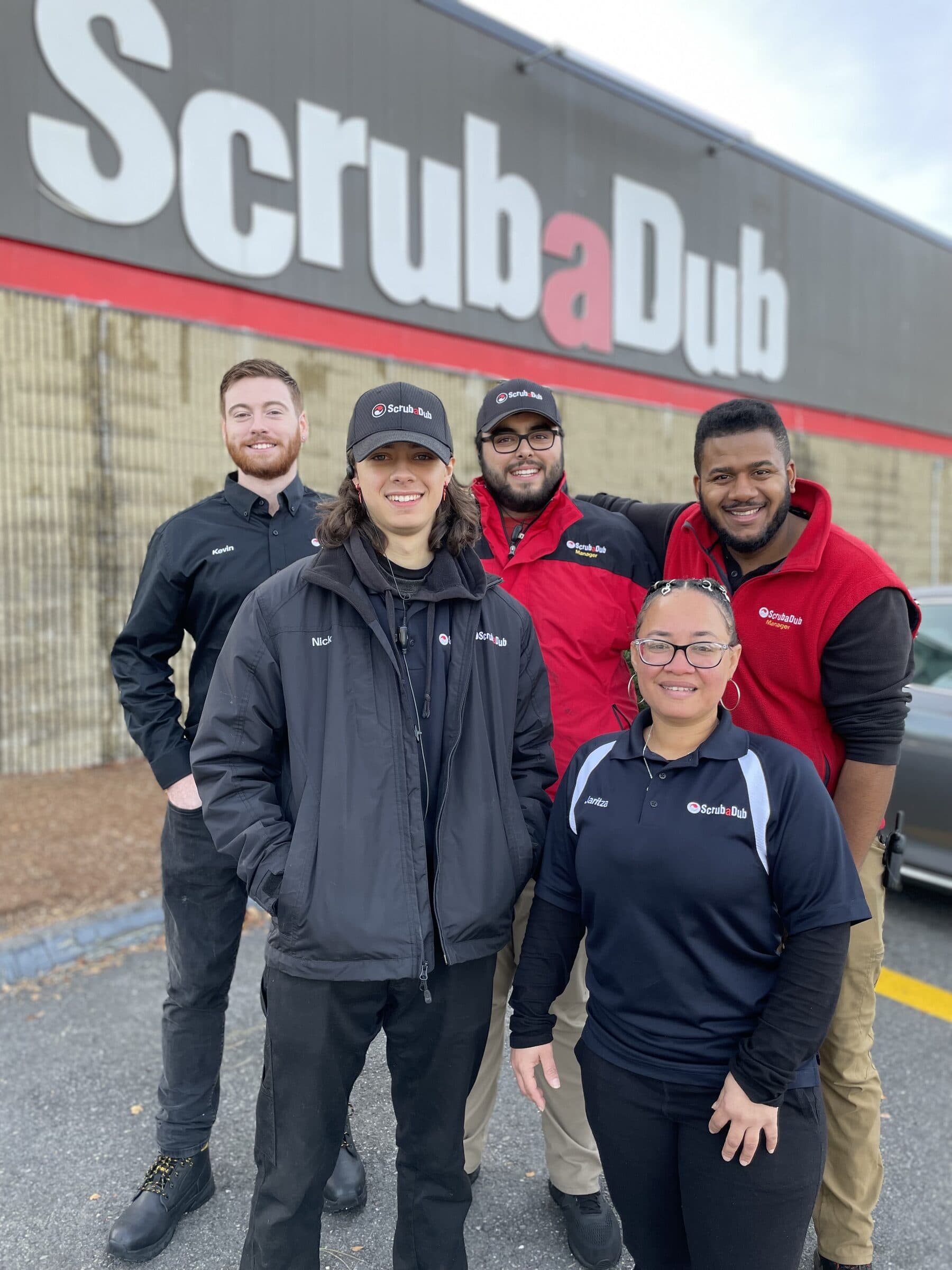 Five smiling ScrubaDub team members in branded jackets and caps standing in front of a large ScrubaDub building sign
