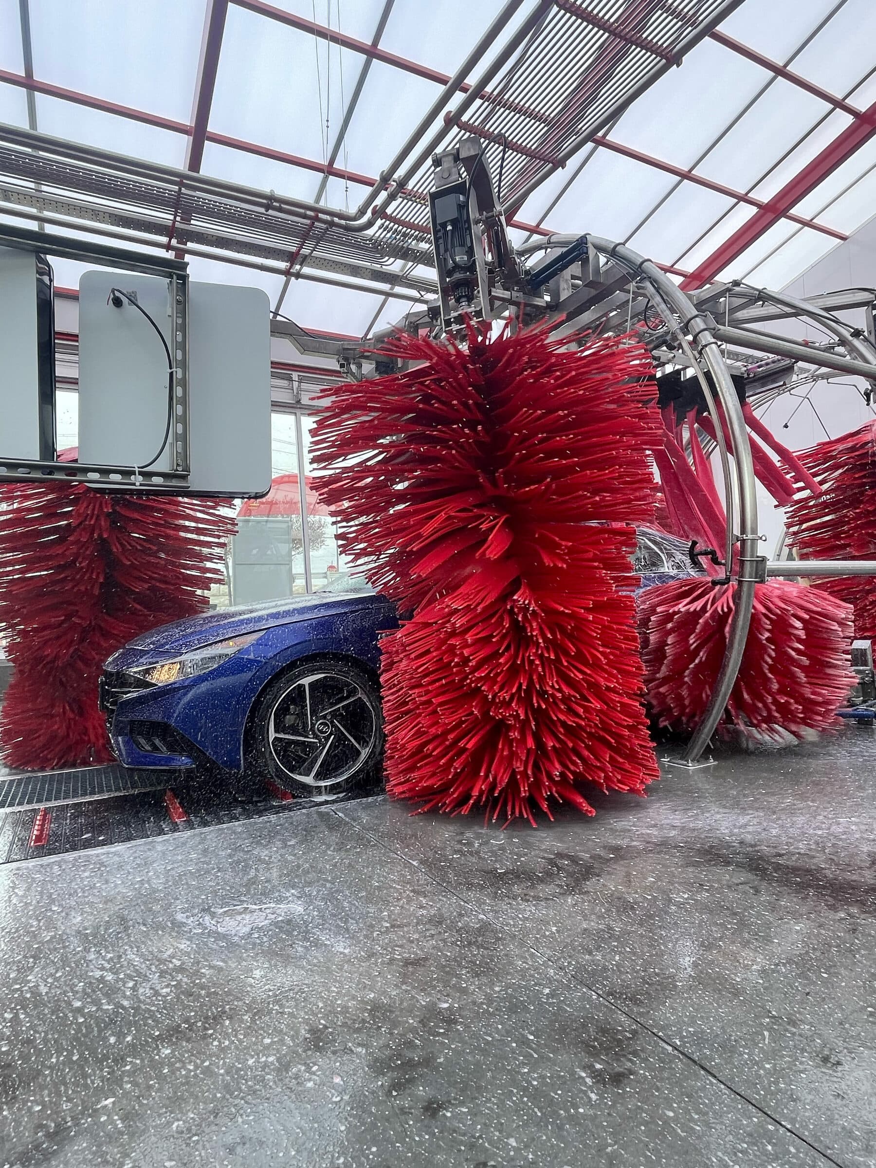 Giant red wash brush spinning beside a blue sedan in a glass-roofed tunnel
