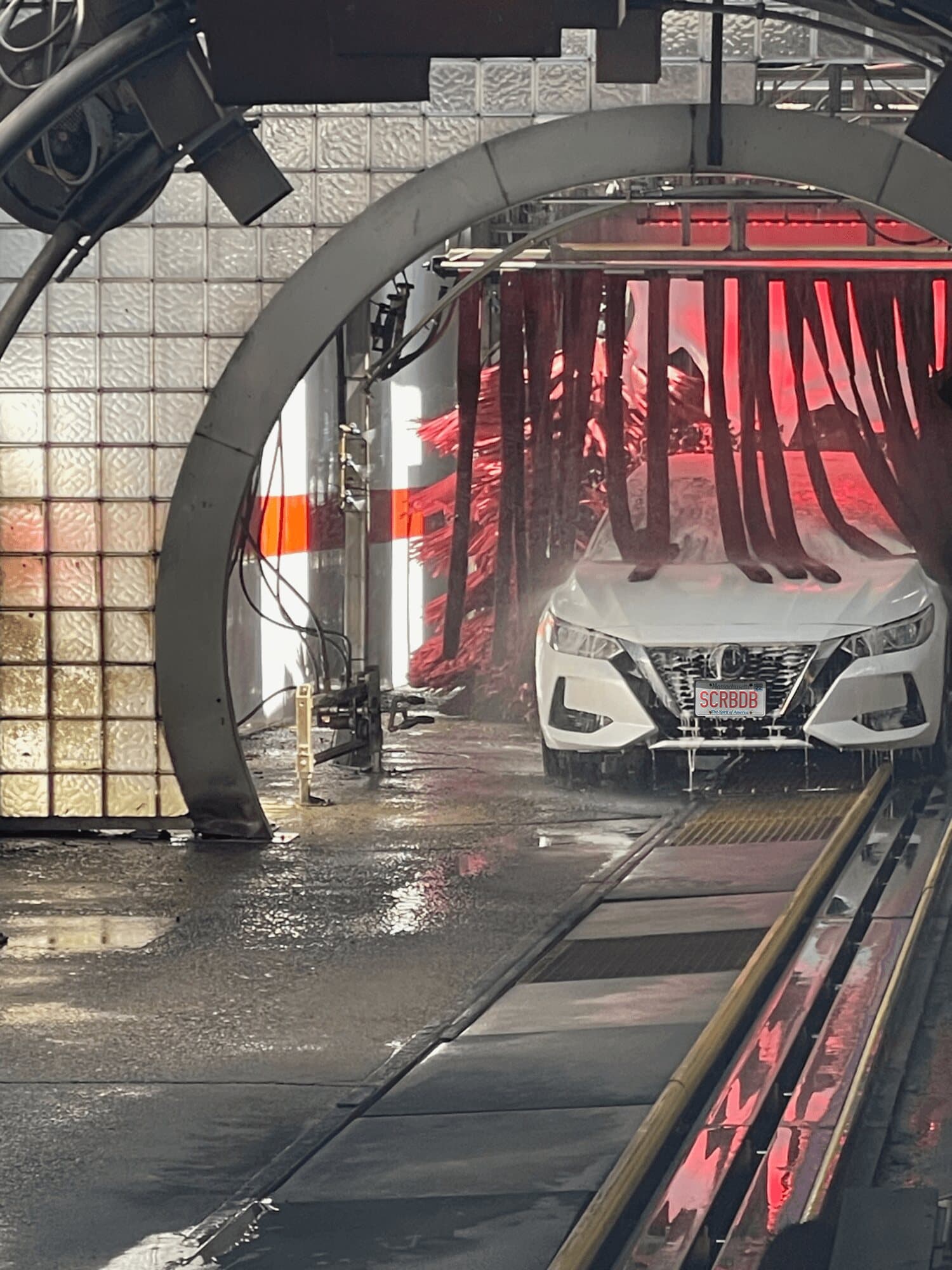 White sedan with a SCRBDB plate moving through red cloth curtains in the wash tunnel
