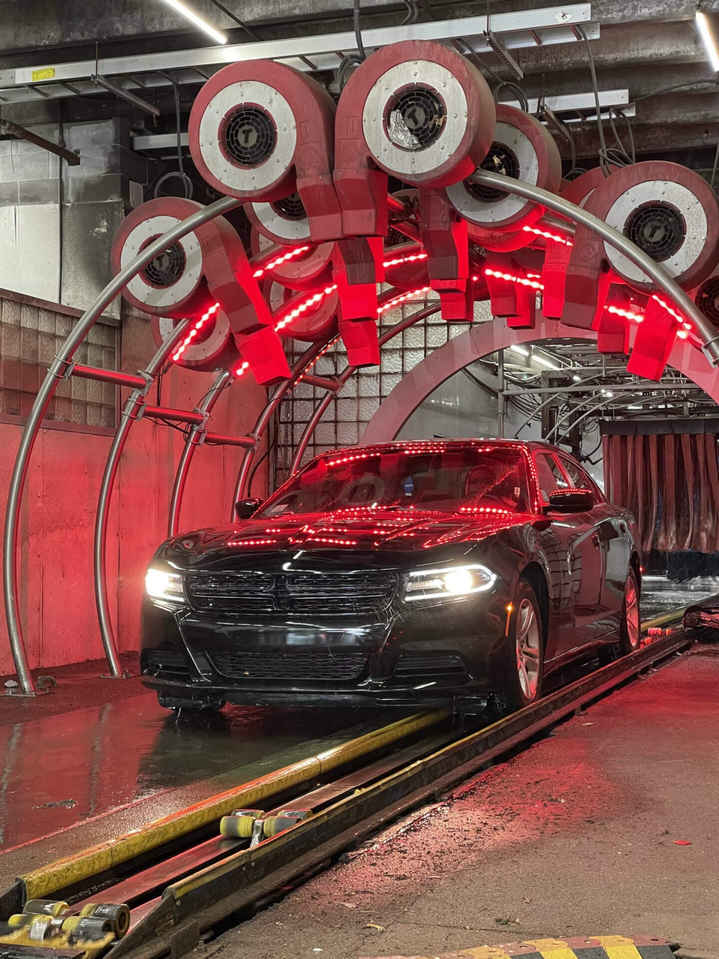Black sedan with headlights on rolling under a red-lit arch of car wash dryers