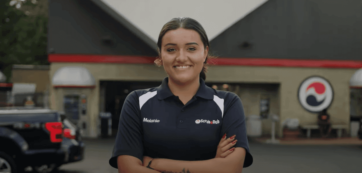 Smiling ScrubaDub employee in a navy uniform polo standing with arms crossed in front of a car wash