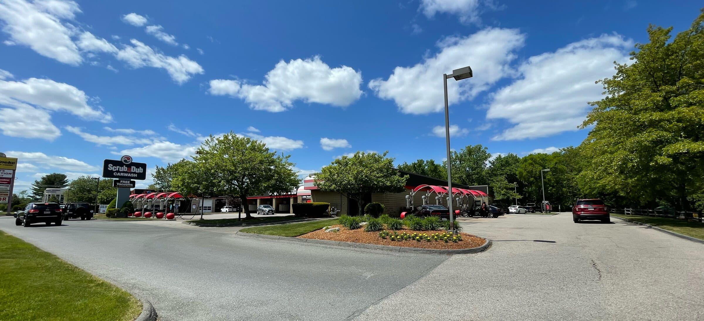 Panoramic view of the ScrubaDub Shrewsbury car wash with its pylon sign, red vacuum canopies and landscaped entrance under a blue sky with white clouds