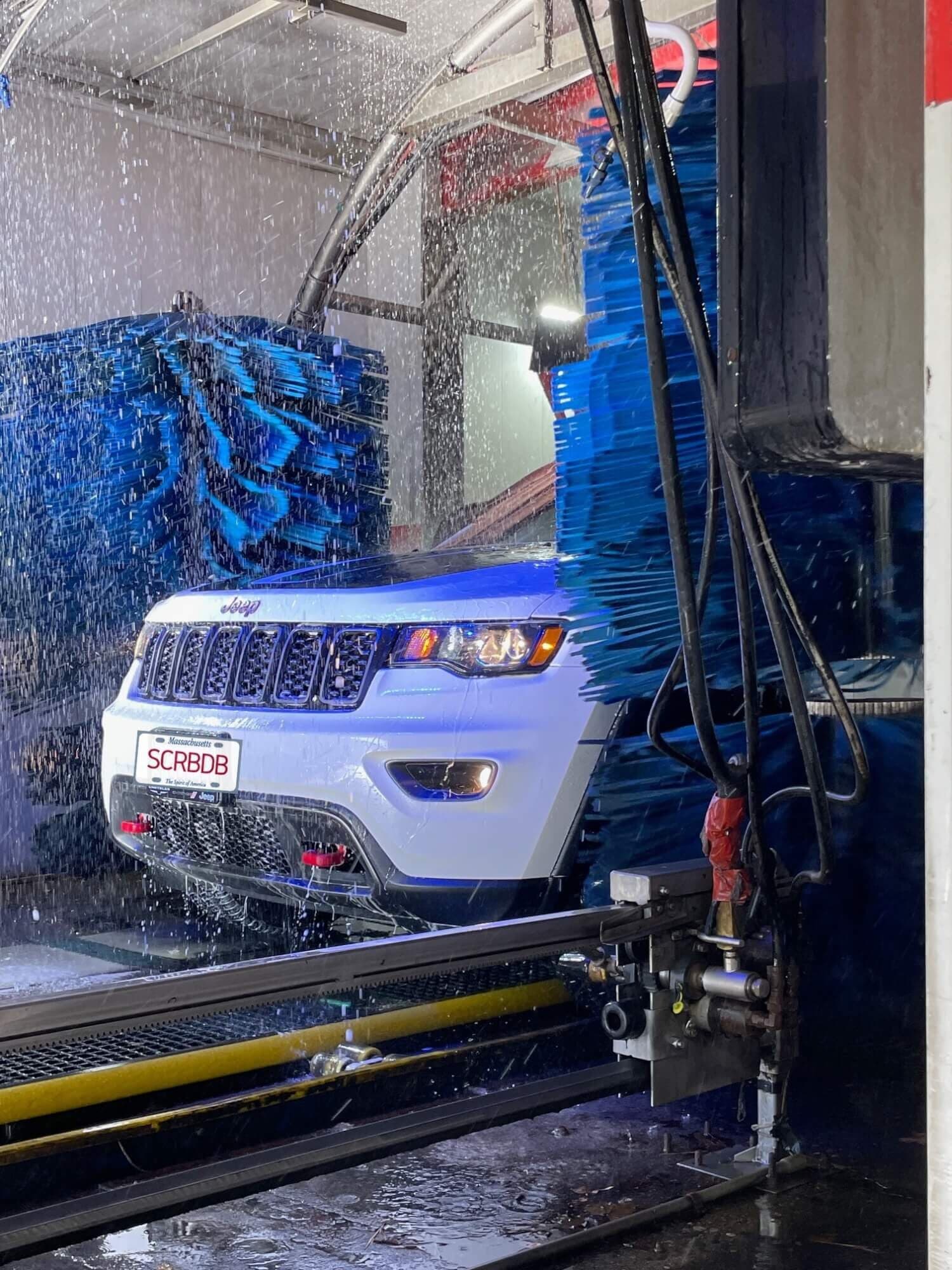 White Jeep with a "SCRBDB" plate moving between spinning blue brushes in a car wash tunnel
