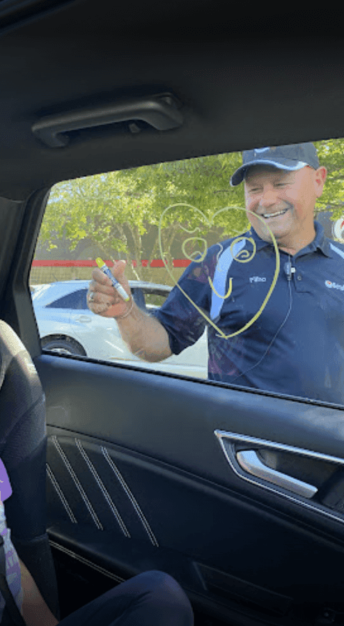 Smiling ScrubaDub attendant drawing a heart on a car window with a marker, seen from the back seat