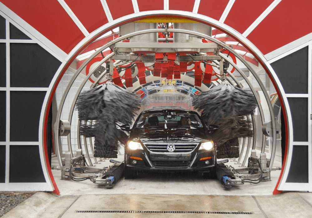 Black Volkswagen sedan with headlights on inside a car wash tunnel entrance framed by a red-and-white arch, wrap brushes spinning on both sides