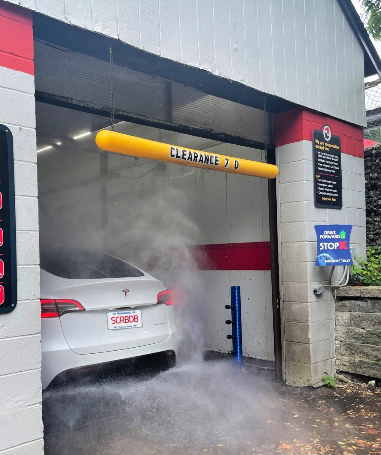 White Tesla with a SCRBDB license plate driving into a misty in-bay car wash under a yellow clearance bar