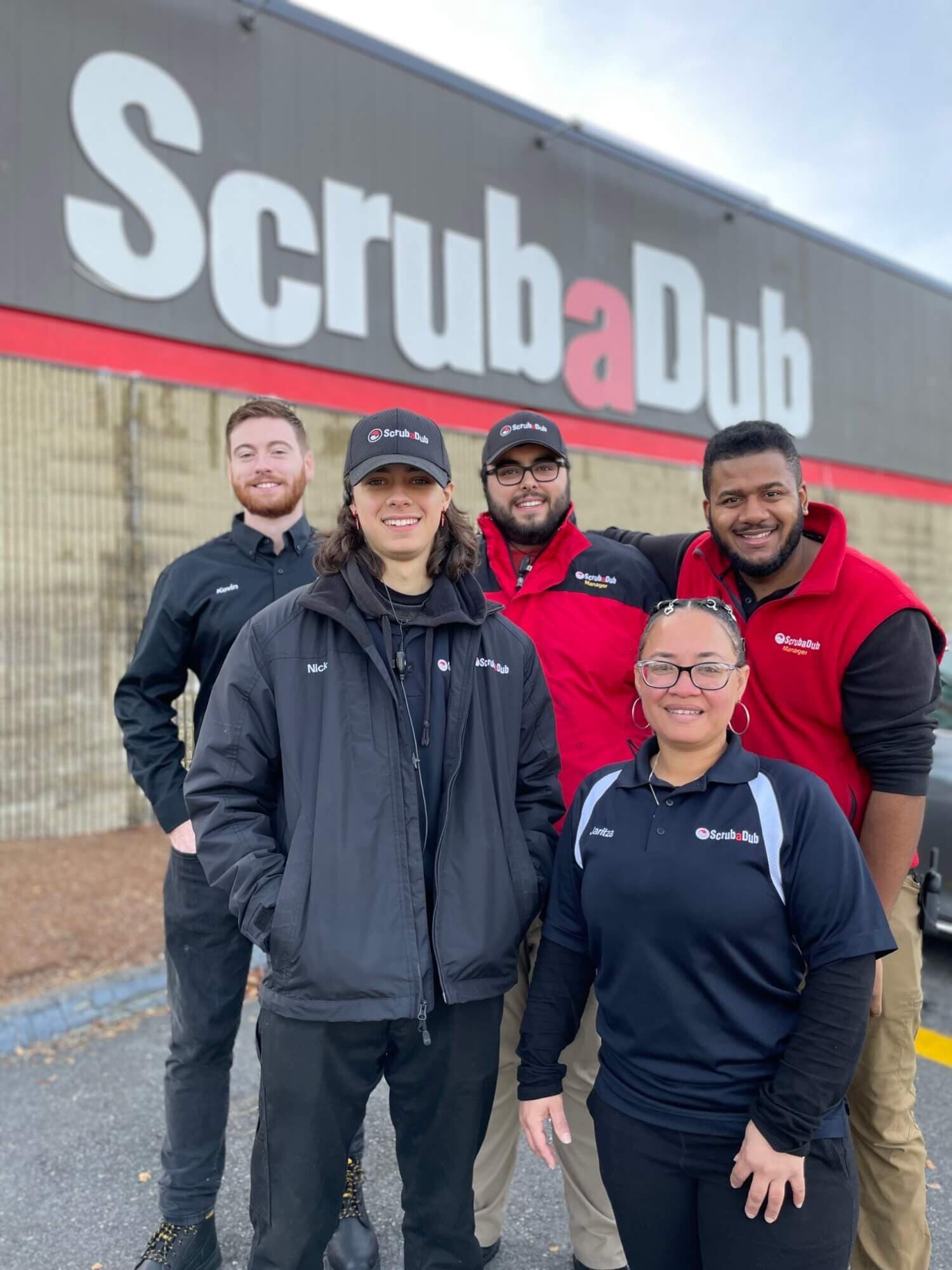 Five smiling ScrubaDub team members in branded jackets and caps standing in front of a large ScrubaDub building sign