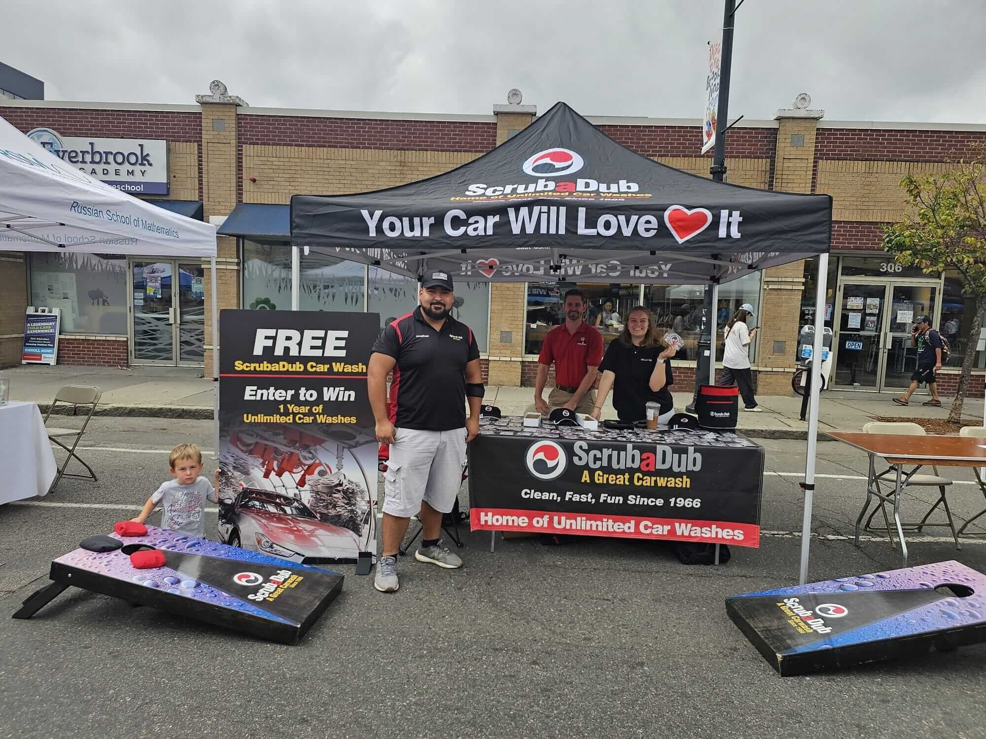 ScrubaDub staff at a street-fair tent reading "Your Car Will Love It" with cornhole boards and an enter-to-win sign