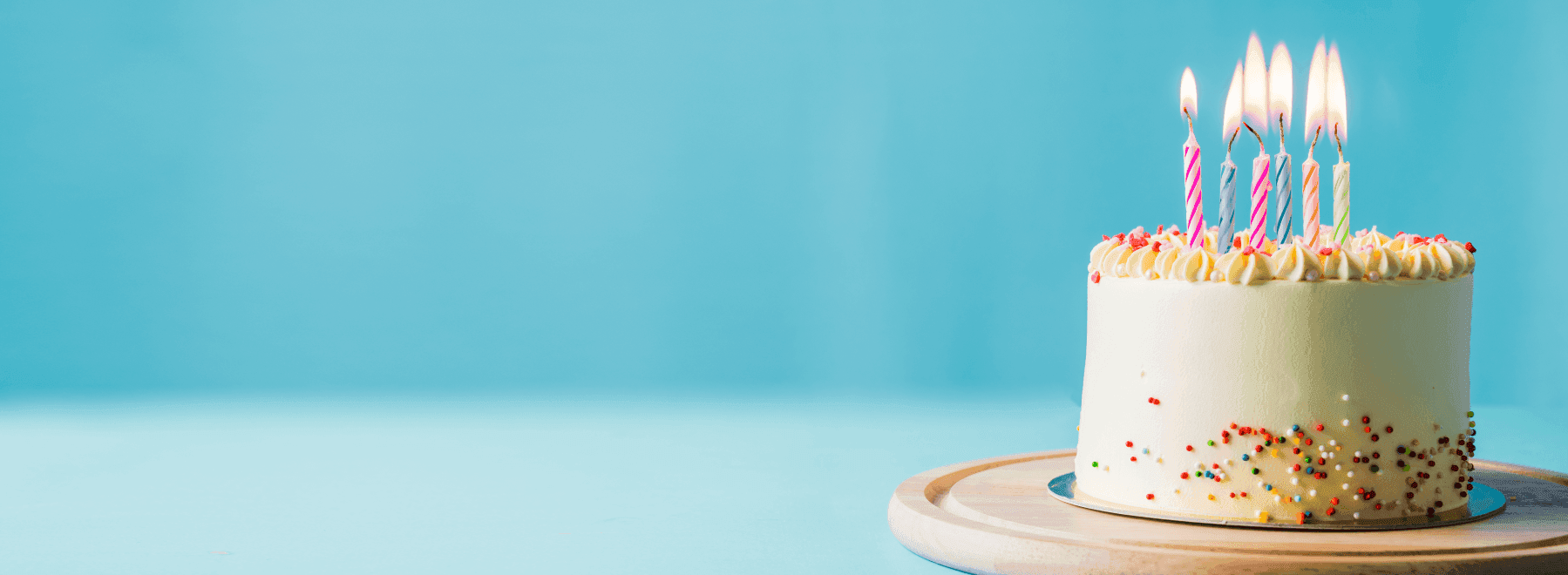 Birthday cake with lit striped candles on a wooden board against a soft blue background