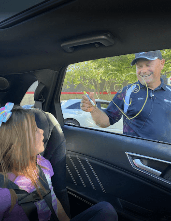 Smiling ScrubaDub attendant drawing a heart on a car window with a marker while a young girl watches from the back seat