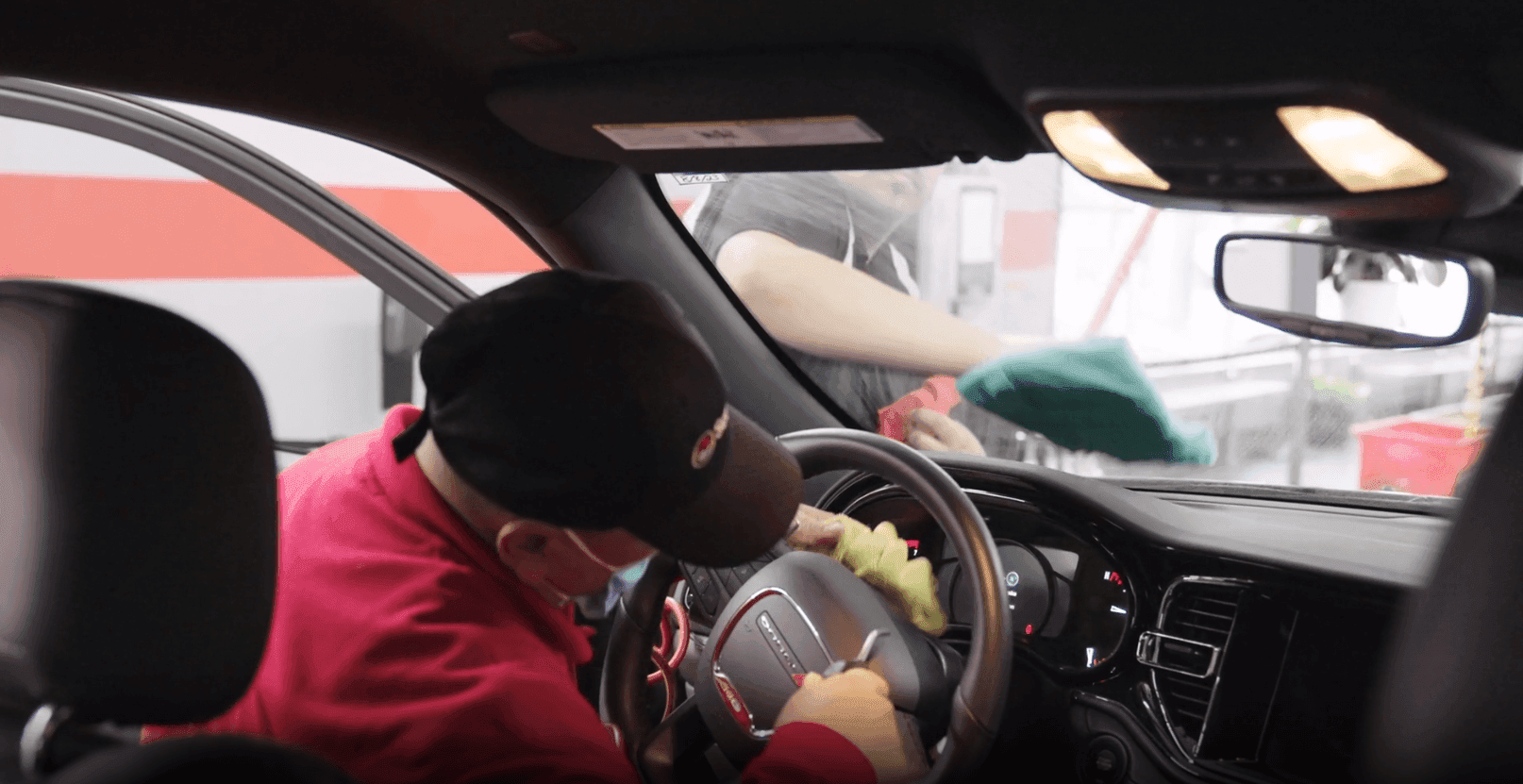 ScrubaDub attendant in a red hoodie and cap wiping a steering wheel while a coworker cleans the windshield from outside