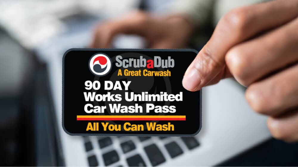 Hand holding a ScrubaDub 90 Day Works Unlimited Car Wash Pass card over a laptop keyboard