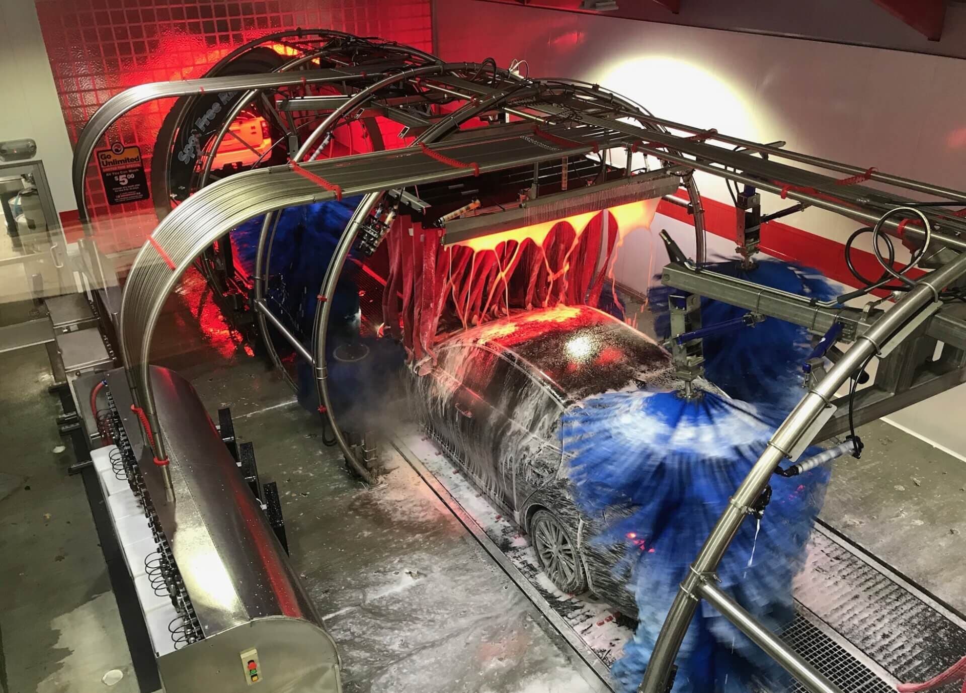 Car covered in soap moving through a tunnel with spinning blue brushes and red-lit cloth curtains