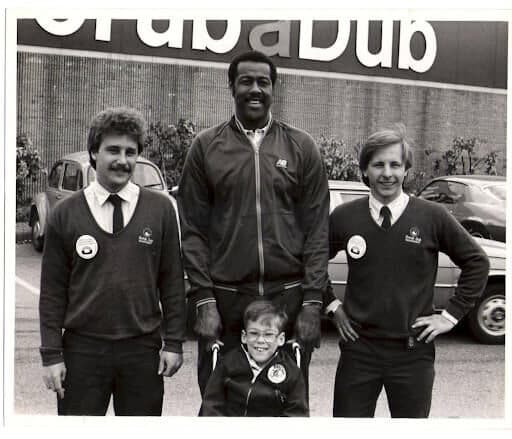 Black-and-white photo of two young ScrubaDub employees in uniform sweaters with a tall man and a small boy in front of a ScrubaDub building