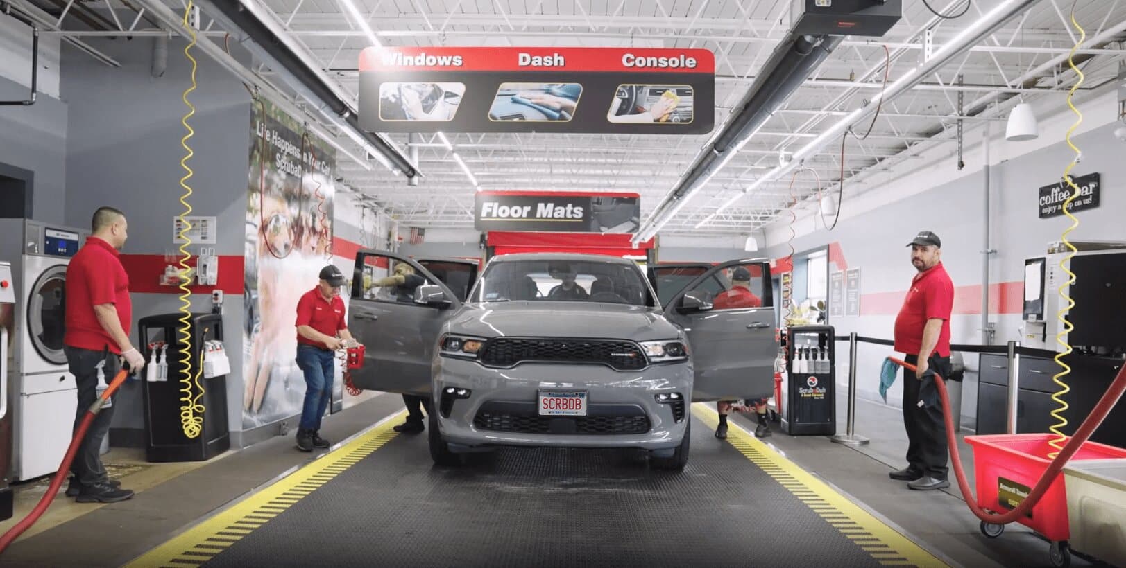 Team of ScrubaDub attendants in red shirts cleaning an SUV inside a bright express interior bay