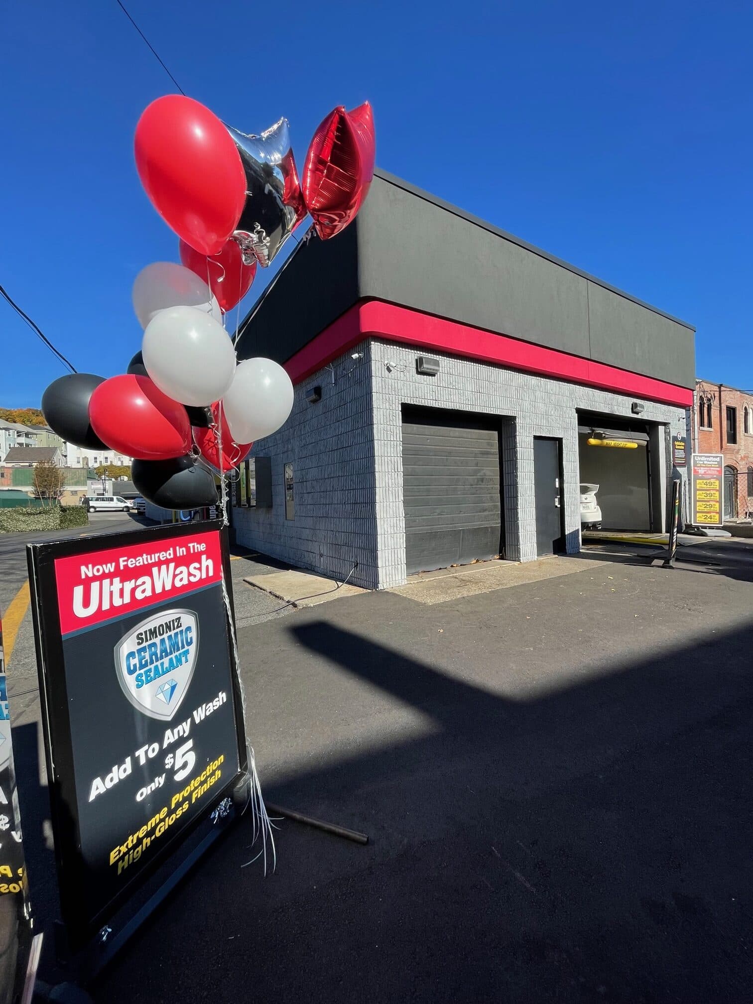 Small ScrubaDub car wash building with red-and-white balloons and an UltraWash ceramic sealant sign under a clear blue sky