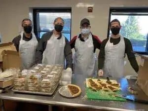 Four masked ScrubaDub volunteers in aprons behind trays of baked goods in a kitchen