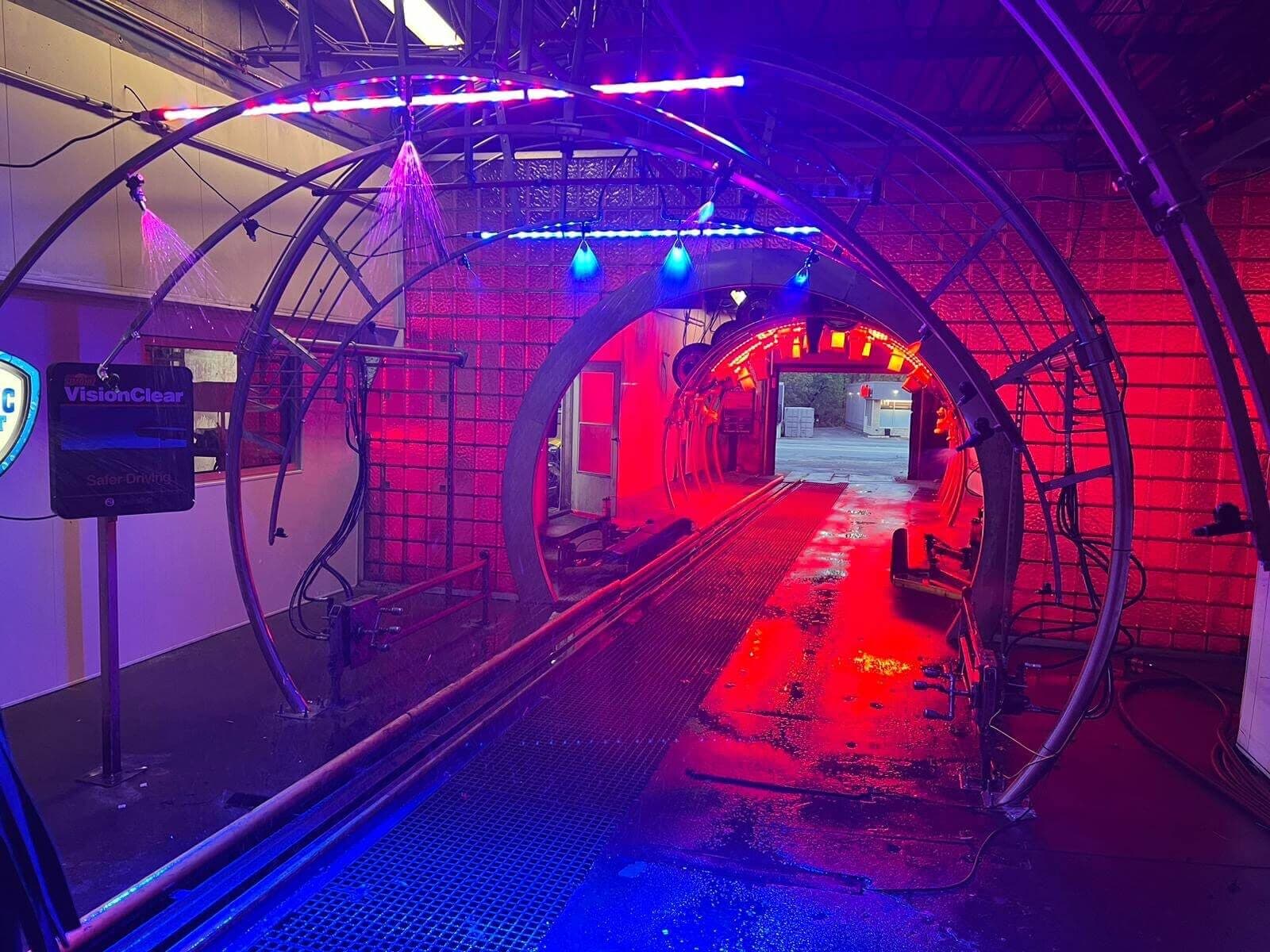 Empty wash tunnel glowing with red and blue lights and stainless arches spraying water
