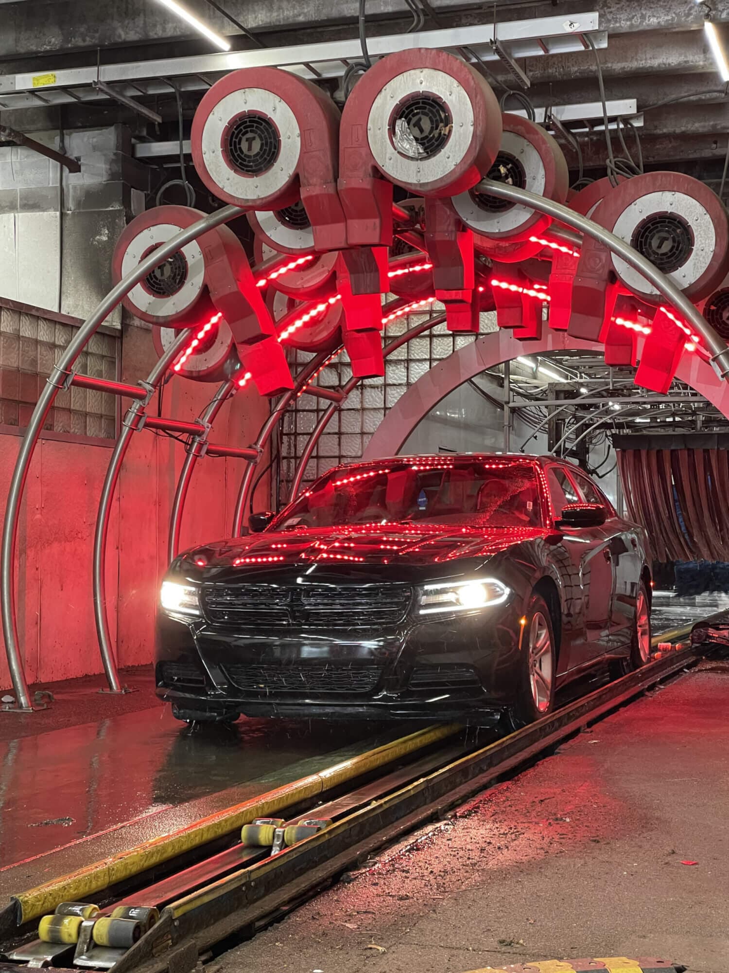 Black sedan with headlights on rolling under a red-lit arch of car wash dryers