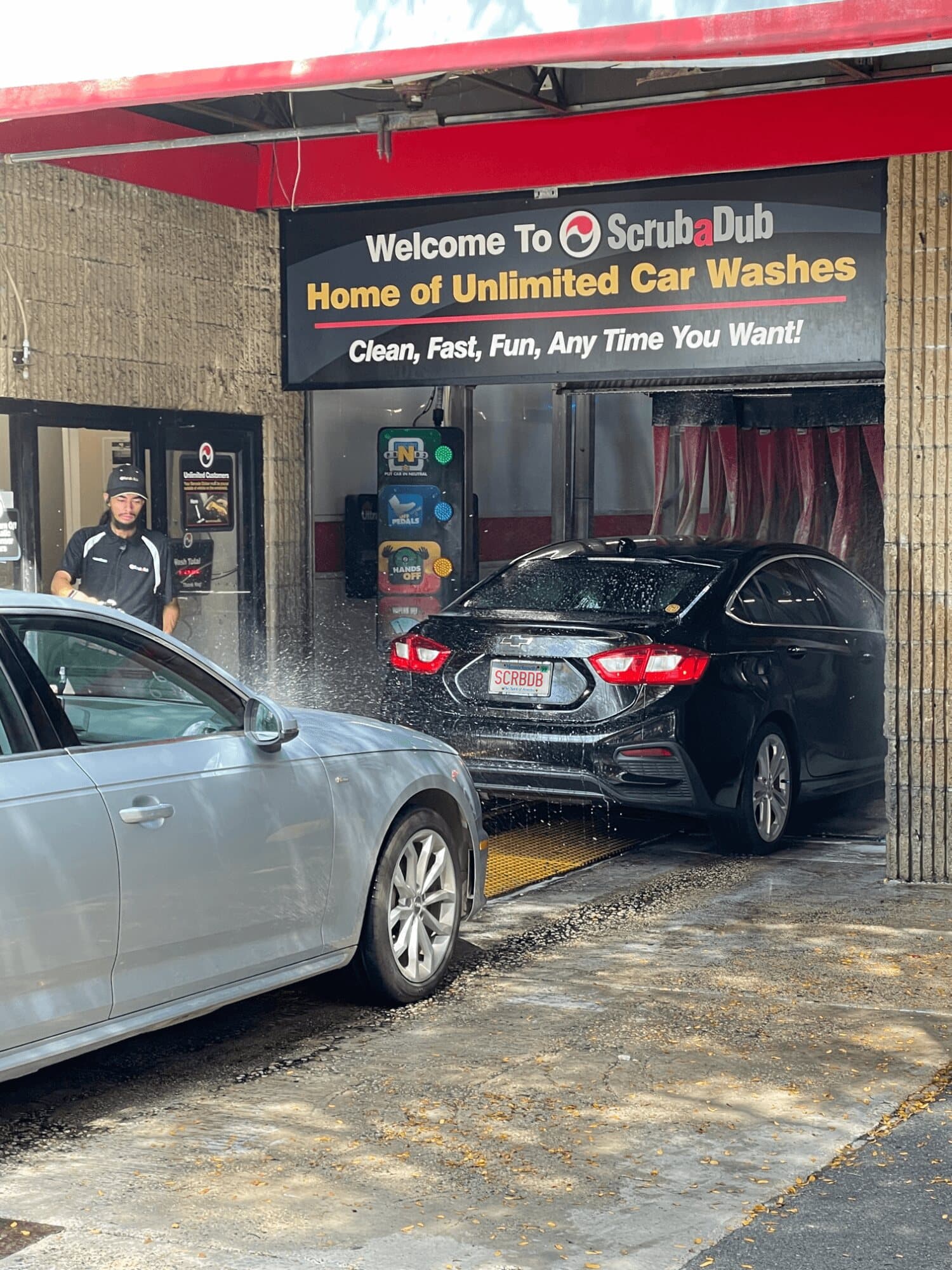 ScrubaDub attendant pre-spraying a silver car as a black sedan enters the tunnel under a Welcome to ScrubaDub sign