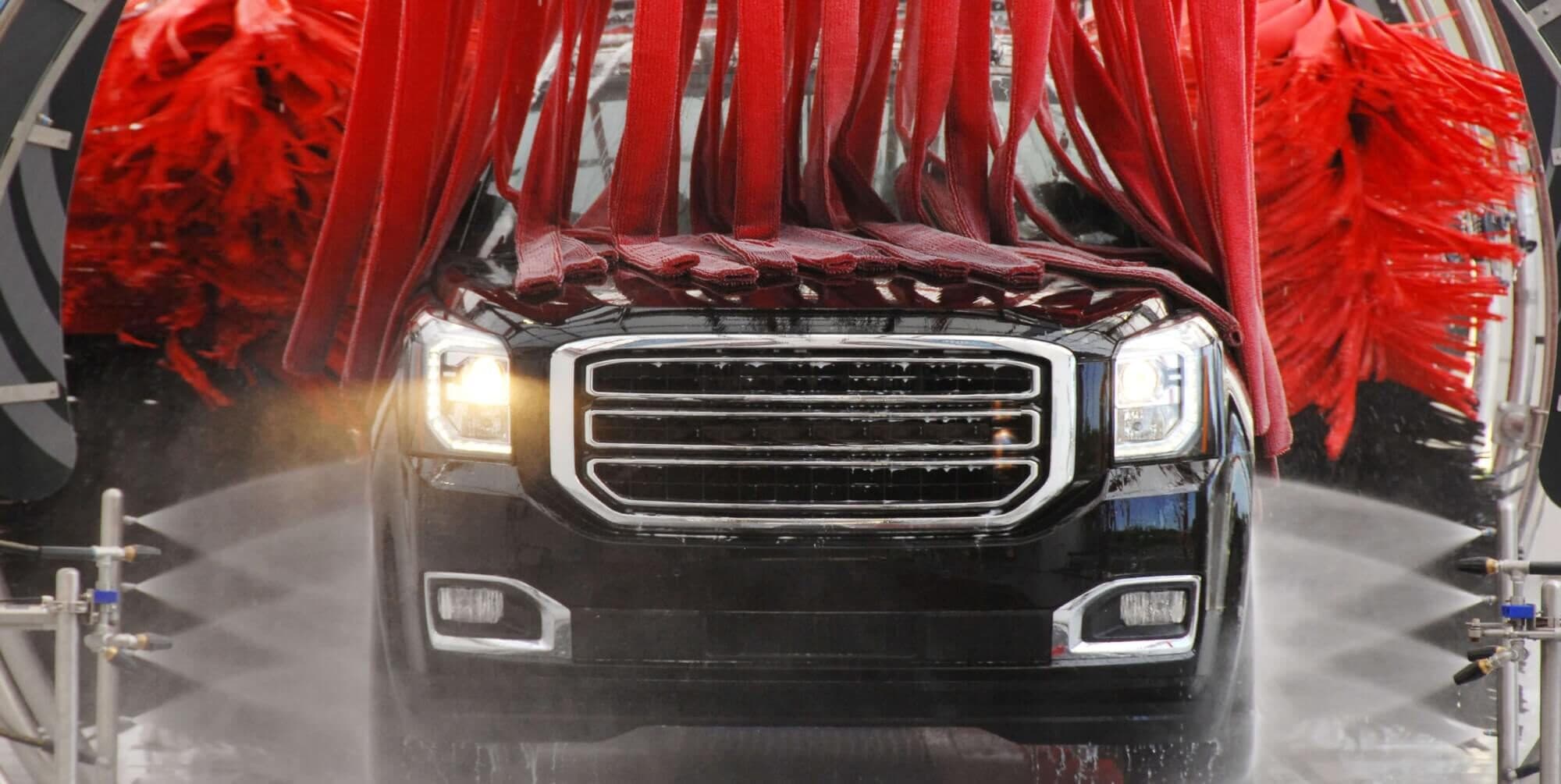 Black GMC SUV with headlights on driving through a car wash tunnel under red cloth strips and spinning red brushes