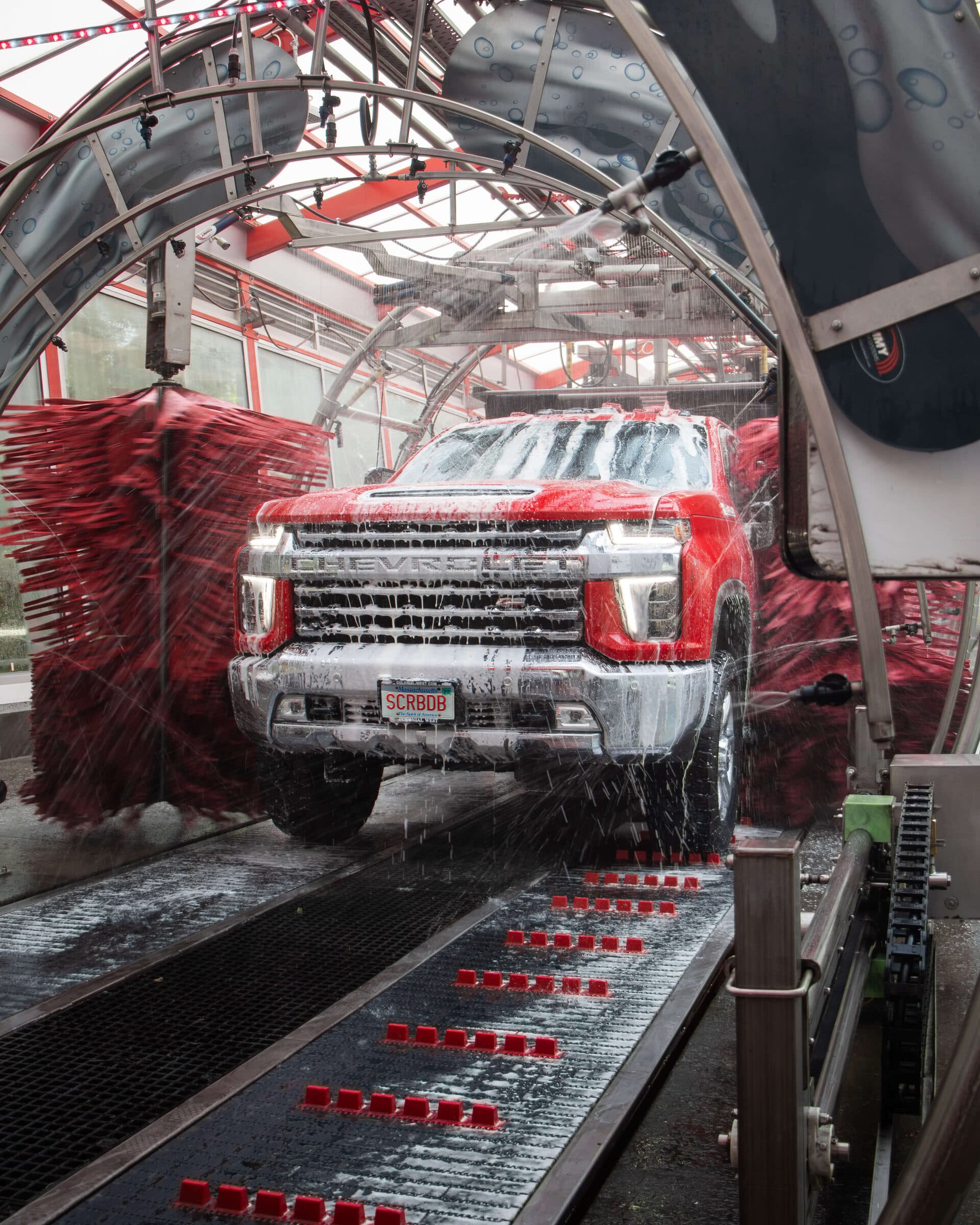 Red Chevrolet pickup covered in soap moving along the conveyor through a car wash tunnel between spinning red brushes