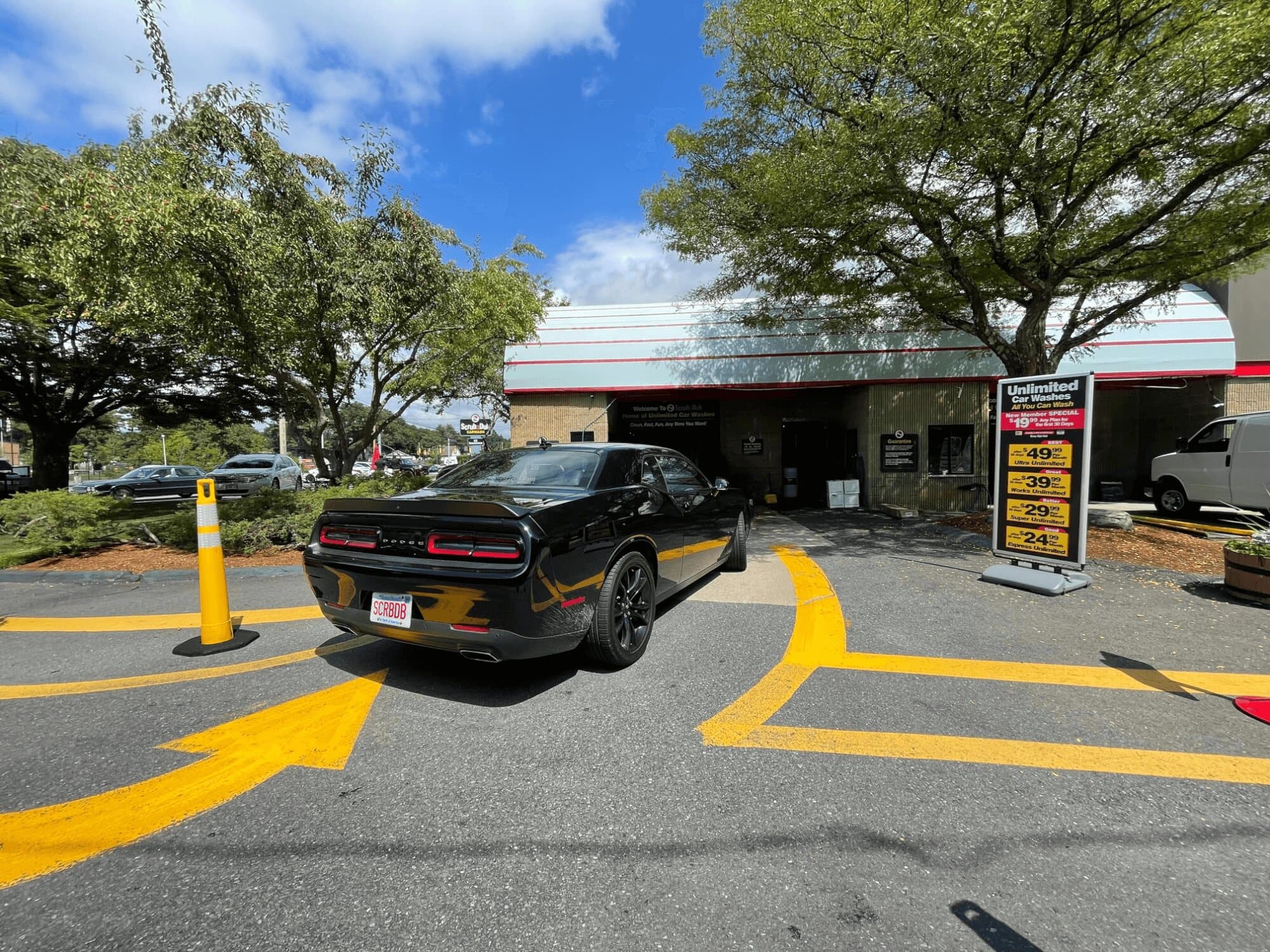 Black muscle car with a SCRBDB plate turning into the ScrubaDub Natick tunnel beside an unlimited pricing sign