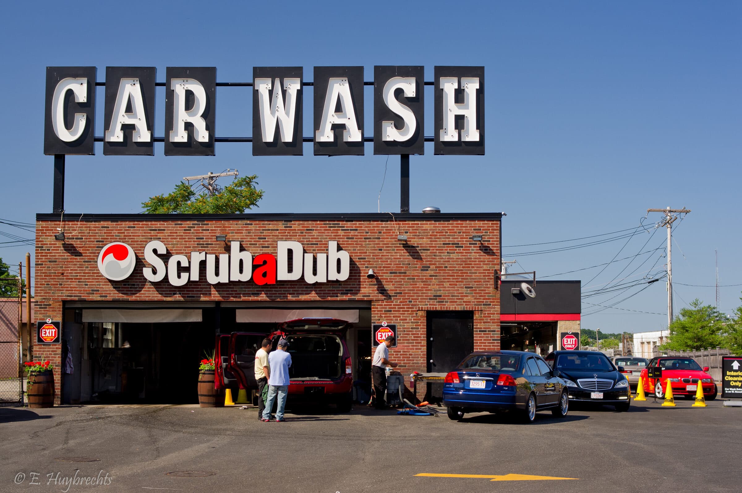 Brick ScrubaDub building under a giant retro CAR WASH rooftop sign, with attendants cleaning a red SUV and cars lined up