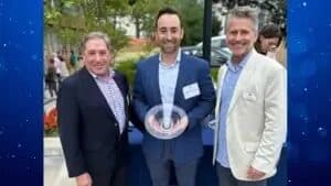 Three men in blazers posing with a glass award at an outdoor evening event